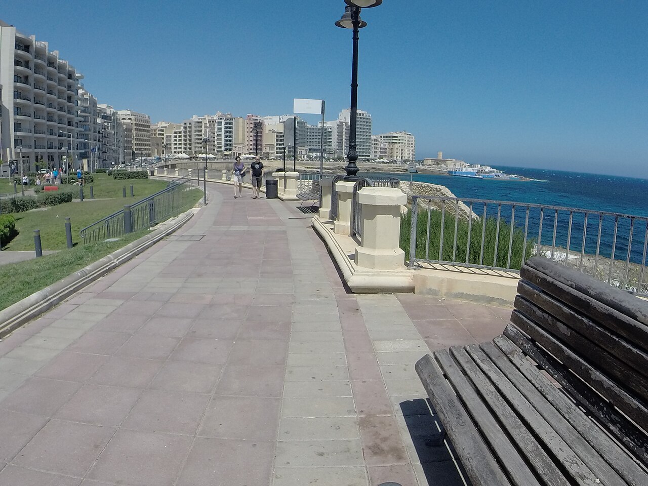 Sliema seafront promenade with a bench, lamps and apartment blocks…