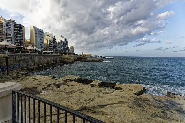 Rocky shoreline at Qui-Si-Sana with waves and seafront buildings under a cloudy sky
